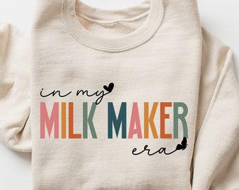 May include: A beige sweatshirt with the text "in my MILK MAKER era" printed in rainbow colors. The text is surrounded by small black hearts.