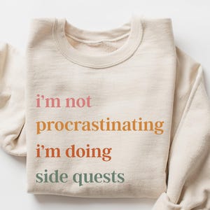 May include: A beige sweatshirt with the text "i'm not procrastinating i'm doing side quests" printed in various colors.