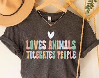 May include: Dark grey heather t-shirt with the text "Loves Animals Tolerates People" in rainbow colors. The shirt is hanging on a wooden hanger with a small heart graphic.