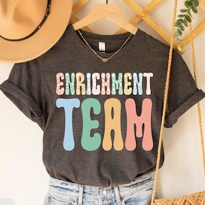 May include: Dark grey heather t-shirt with the text "Enrichment Team" in colorful, retro-style lettering. The shirt is being modeled on a person wearing blue jeans and a brown hat.