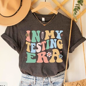May include: Dark grey heather t-shirt with the text "In My Testing Era" in colorful retro font with a pencil and star accents.
