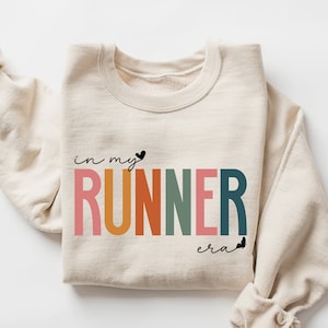 May include: A beige sweatshirt with the text "in my runner era" in rainbow colors and black hearts.