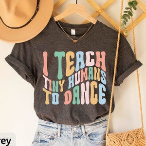 May include: Dark grey heather t-shirt with the text "I teach tiny humans to dance" in colorful, retro-style lettering.