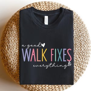 May include: A black crew neck t-shirt featuring the white cursive text 'a good' followed by a white heart, the colorful block text 'WALK FIXES' in lavender, pink, yellow, teal, and purple, and the white cursive text 'everything' with a butterfly.