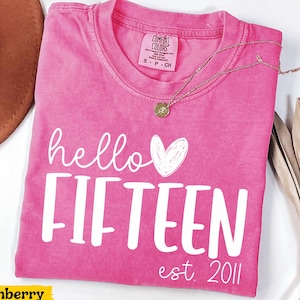 May include: A pink short-sleeved t-shirt with a white graphic print that reads hello FIFTEEN est. 2011 and includes a hand-drawn heart. The shirt features a crew neckline and a Comfort Colors brand label on the inside collar area.