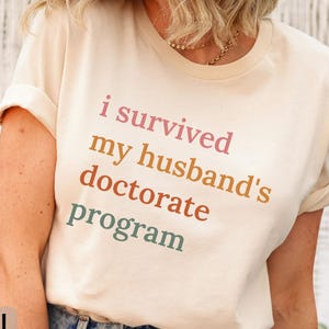 May include: A natural-colored t-shirt with the text "i survived my husband's doctorate program" in pink, orange, and green. The shirt is made of Bella Canvas 3001 material.