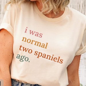 May include: A natural-colored Bella Canvas 3001 t-shirt with the text "i was normal two spaniels ago." The text is in a gradient of pink, orange, and green. The shirt has rolled-up sleeves and is paired with blue jeans.