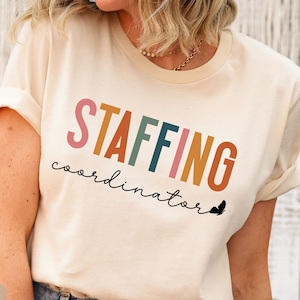 May include: A beige Bella Canvas 3001 t-shirt with the text "STAFFING coordinator" in rainbow colors. The text is in a stylized font with a small butterfly graphic below it.