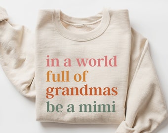 May include: A beige sweatshirt with the text "in a world full of grandmas be a mimi" printed in rainbow colors.