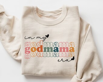 May include: A beige sweatshirt with the text "in my godmama era" printed on the front. The text is in a rainbow colour scheme with a heart at the end.