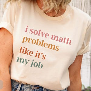 May include: A natural-colored Bella Canvas 3001 t-shirt with the text "i solve math problems like it's my job" in pink, orange, and green. The shirt has rolled-up sleeves and is paired with blue jeans.
