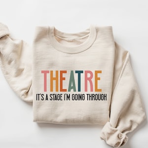 May include: A beige sweatshirt with the text "THEATRE IT'S A STAGE I'M GOING THROUGH" printed on the front. The word "THEATRE" is in colorful letters.