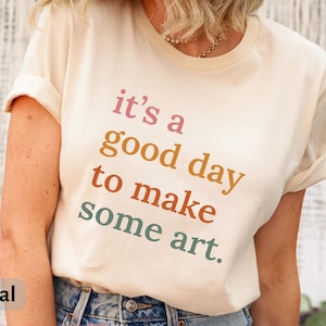 May include: A cream-colored t-shirt with the text "it's a good day to make some art" in a gradient of pink, orange, red, and green. The shirt is a Bella Canvas 3001, and the color is natural.