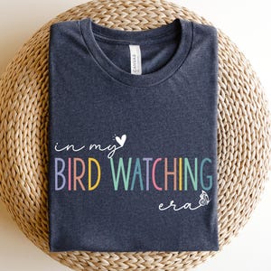 May include: Heather navy t-shirt with the text "in my bird watching era" in colorful letters. The shirt has a crew neck and is made of a soft material. The shirt is displayed on a woven, circular surface.