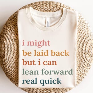 May include: A cream-colored t-shirt with the text "i might be laid back but i can lean forward real quick" in pink, orange, rust, teal, and navy blue. The word "natural" is in a yellow rectangle in the lower left corner. The shirt is on a woven surface.