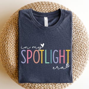 May include: Heather navy t-shirt with the text "in my SPOTLIGHT era" in colorful letters. The design includes a white heart and butterfly. The shirt is displayed on a woven, circular surface.