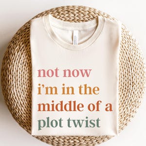 May include: A white t-shirt with the text "not now i'm in the middle of a plot twist" printed in pink, orange, brown, and green.