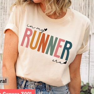 Peut inclure: Un t-shirt beige avec le texte "in my runner era" en couleurs arc-en-ciel. Le texte est dans un style de police rétro avec un petit symbole de cœur après "my".