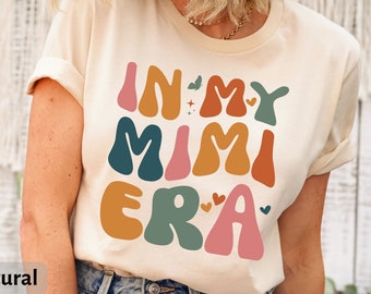Pode incluir: Uma camiseta bege com o texto "In My Mimi Era" em letras coloridas de estilo retrô. As letras são em tons de laranja, rosa, verde e azul.