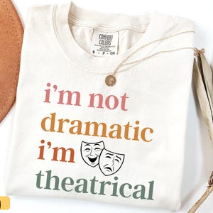 May include: Ivory-colored Comfort Colors t-shirt with the text "I'm not dramatic I'm theatrical" in pink, orange, and green. Features two theater masks. A gold necklace and a brown hat are also visible.
