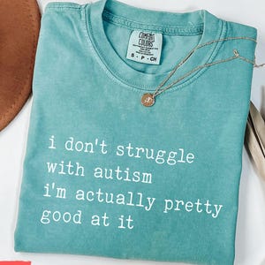 May include: Seafoam green t-shirt with the text "i don't struggle with autism i'm actually pretty good at it" in white. A gold necklace with a pendant is on the shirt. The shirt is folded on a white surface.