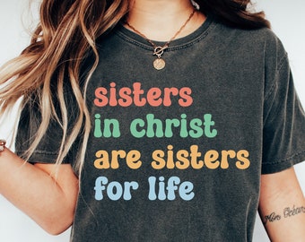 May include: A dark gray t-shirt with the text "sisters in christ are sisters for life" printed in colorful letters. The text is in a retro font style.