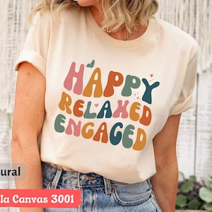 May include: A beige t-shirt with the text "Happy Relaxed Engaged" in colorful, retro-style lettering. The shirt is made of Bella Canvas 3001 fabric and is available in natural color.