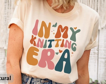 May include: A beige t-shirt with the text "In My Knitting Era" in colorful, retro-style lettering. The shirt is made by Bella Canvas and is a 3001 style.