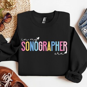 Peut inclure: Sweat-shirt noir avec le texte "in my sonographer era" en couleurs arc-en-ciel avec un petit cœur.