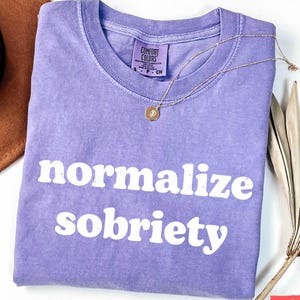 May include: A violet Comfort Colors t-shirt with the words "normalize sobriety" in white block letters. A gold necklace with a pendant rests on the shirt. A brown hat and dried plant are in the background.