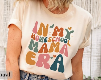 Może przedstawiać: Beżowa koszulka z napisem "In My Homeschool Mama Era" w kolorowych, retro literach. Koszulkę nosi osoba z długimi brązowymi włosami.