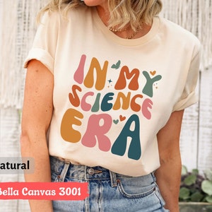 May include: A beige t-shirt with the text "In My Science Era" in colorful, retro-style lettering. The shirt is made of Bella Canvas 3001 fabric and is available in natural color.