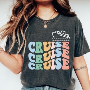 May include: A dark gray t-shirt with a white outline of a cruise ship and the word "CRUISE" repeated three times in colorful, retro-style lettering.