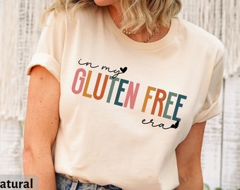 May include: A beige t-shirt with the text "in my gluten free era" in rainbow colors. The text is in a playful font with a small heart and a butterfly graphic. The shirt is made of Bella Canvas 3001 fabric and is available in natural color.