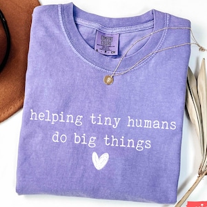 May include: A violet Comfort Colors t-shirt with the text "helping tiny humans do big things" and a small white heart. A gold necklace and a brown hat are also in the image.