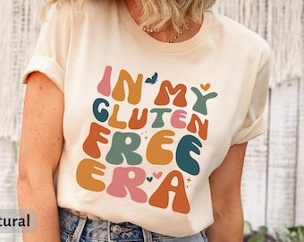 May include: A beige t-shirt with the text "In My Gluten Free Era" in colorful, retro-style lettering. The shirt is being worn by a person with their arms at their sides.