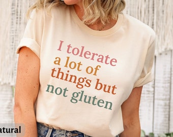 May include: A beige t-shirt with the text "I tolerate a lot of things but not gluten" printed in various colors. The text is in a modern font style.