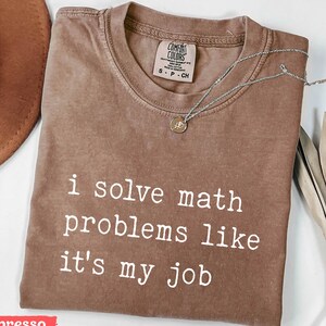 May include: Espresso-colored t-shirt with the text "i solve math problems like it's my job" in white. The shirt has a crew neck and short sleeves. A silver necklace with a small pendant is draped across the shirt.