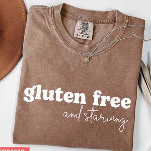 Puede incluir: Camiseta color espresso con las palabras "gluten free and starving" en escritura blanca. Un collar plateado con un peque&ntilde;o colgante descansa sobre la camiseta. La camiseta es de la marca Comfort Colors.