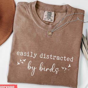May include: Espresso-colored t-shirt with the phrase "easily distracted by birds" in white script. The shirt has a crew neck and short sleeves. A silver necklace with a small pendant rests on the shirt.