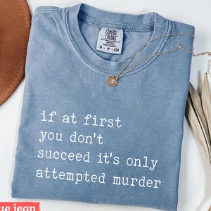 May include: A blue jean colored t-shirt with the text "if at first you don't succeed it's only attempted murder" in white. A gold necklace with a pendant is draped across the shirt.