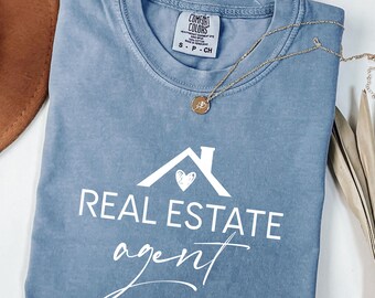 May include: A blue jean t-shirt with a white graphic of a house with a heart inside and the text "REAL ESTATE agent" in a cursive font.