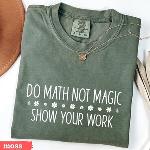 May include: A green t-shirt with the text "Do Math Not Magic Show Your Work" in white. The shirt has a row of white daisies below the text. The shirt is on a white surface.