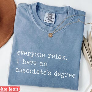 May include: Blue jean colored t-shirt with the text "everyone relax, I have an associate's degree" in white. A gold necklace with a pendant is draped across the shirt. The shirt is folded and has a Comfort Colors tag.