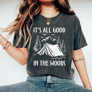 May include: A dark gray t-shirt with a white graphic of a tent in the woods. The text "It's all good in the woods" is printed on the shirt.
