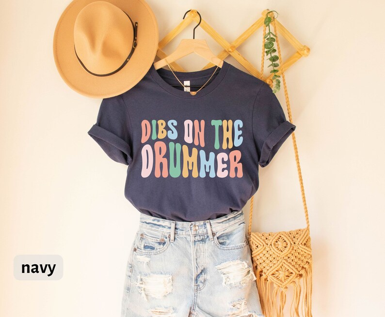 Puede incluir: Una camiseta azul marino con el texto "Dibs on the Drummer" en colores arco&iacute;ris. La camiseta se muestra en una percha con un sombrero de paja y una bolsa tejida.