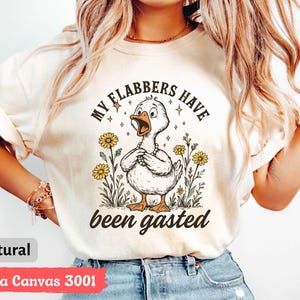 May include: A natural-colored t-shirt with the text "MY FLABBERS HAVE been gasted" and a cartoon duck wearing a pearl necklace. The duck is surrounded by yellow flowers and greenery. The shirt is labeled "Bella Canvas 3001".
