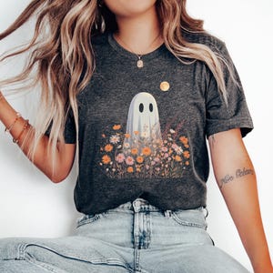 Pu&ograve; includere: T-shirt grigio scuro con un design a fantasma in un campo di fiori colorati. Il fantasma ha due occhi ovali neri ed &egrave; circondato da fiori arancioni, rosa e gialli. Una luna dorata &egrave; nell'angolo in alto a destra.
