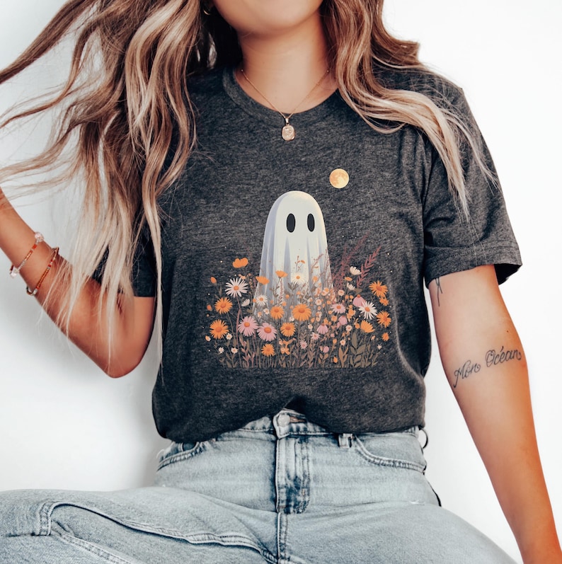 Pu&ograve; includere: T-shirt grigio scuro con un design a fantasma in un campo di fiori colorati. Il fantasma ha due occhi ovali neri ed &egrave; circondato da fiori arancioni, rosa e gialli. Una luna dorata &egrave; nell'angolo in alto a destra.