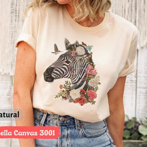 May include: A natural colored t-shirt with a zebra print design. The zebra is surrounded by flowers and birds. The text "Bella Canvas 3001" is printed on the bottom of the shirt.